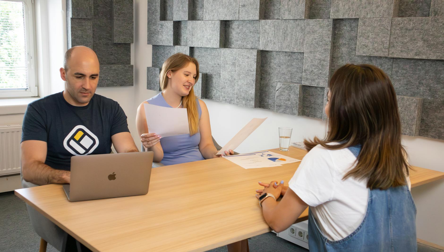 Three colleagues in a modern office discussing work with laptops and documents.