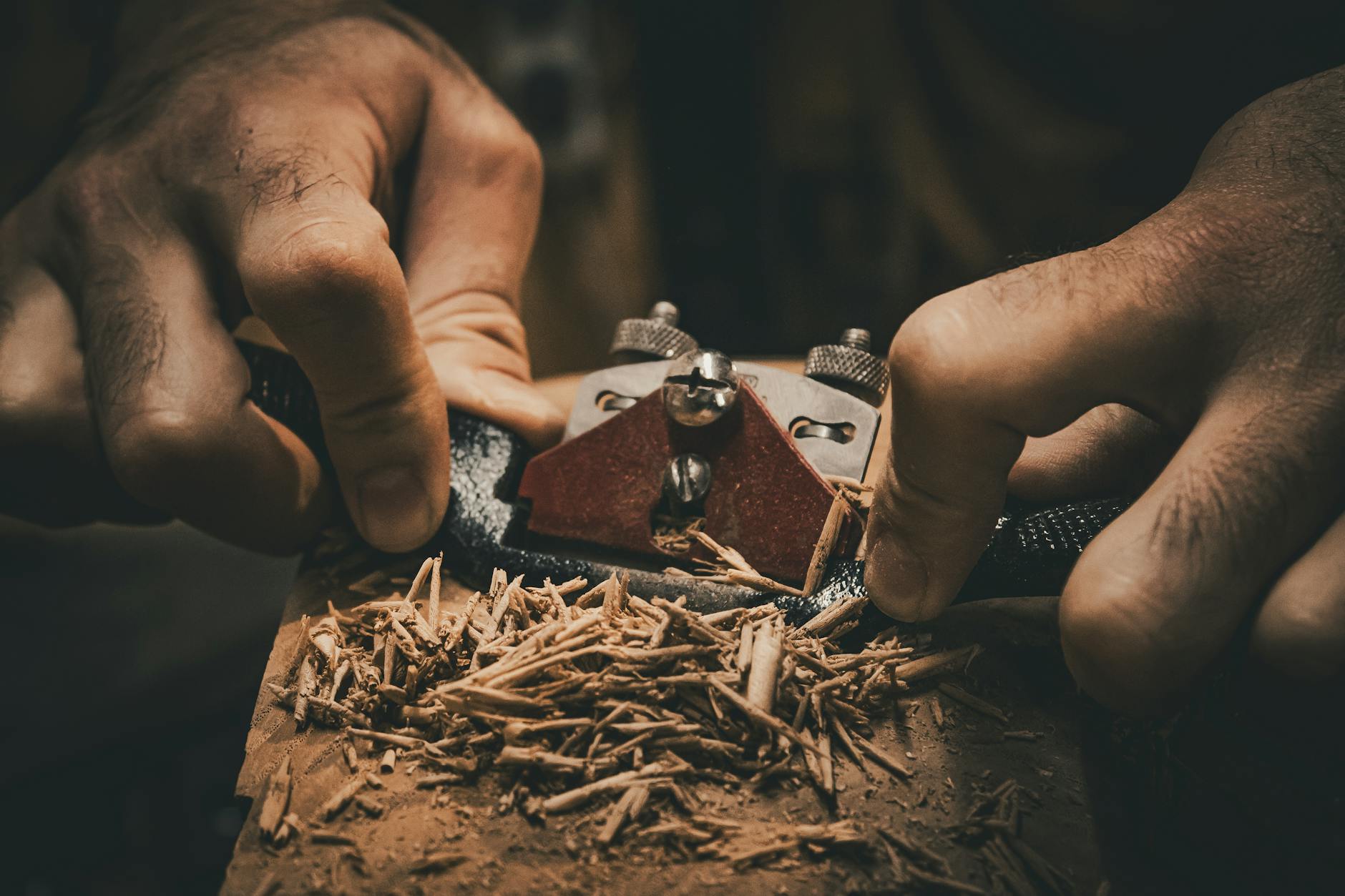 Detailed view of hands using a hand plane tool in woodworking, creating wood shavings.