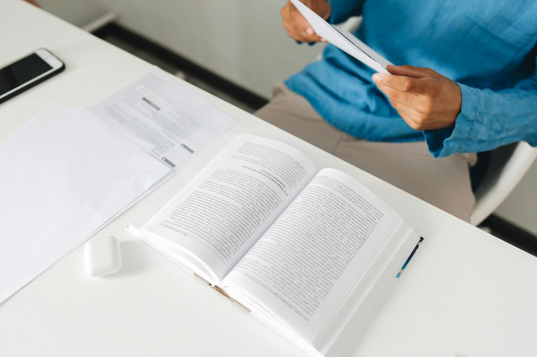 An open book on a desk with a person reading documents beside it in an office setting.