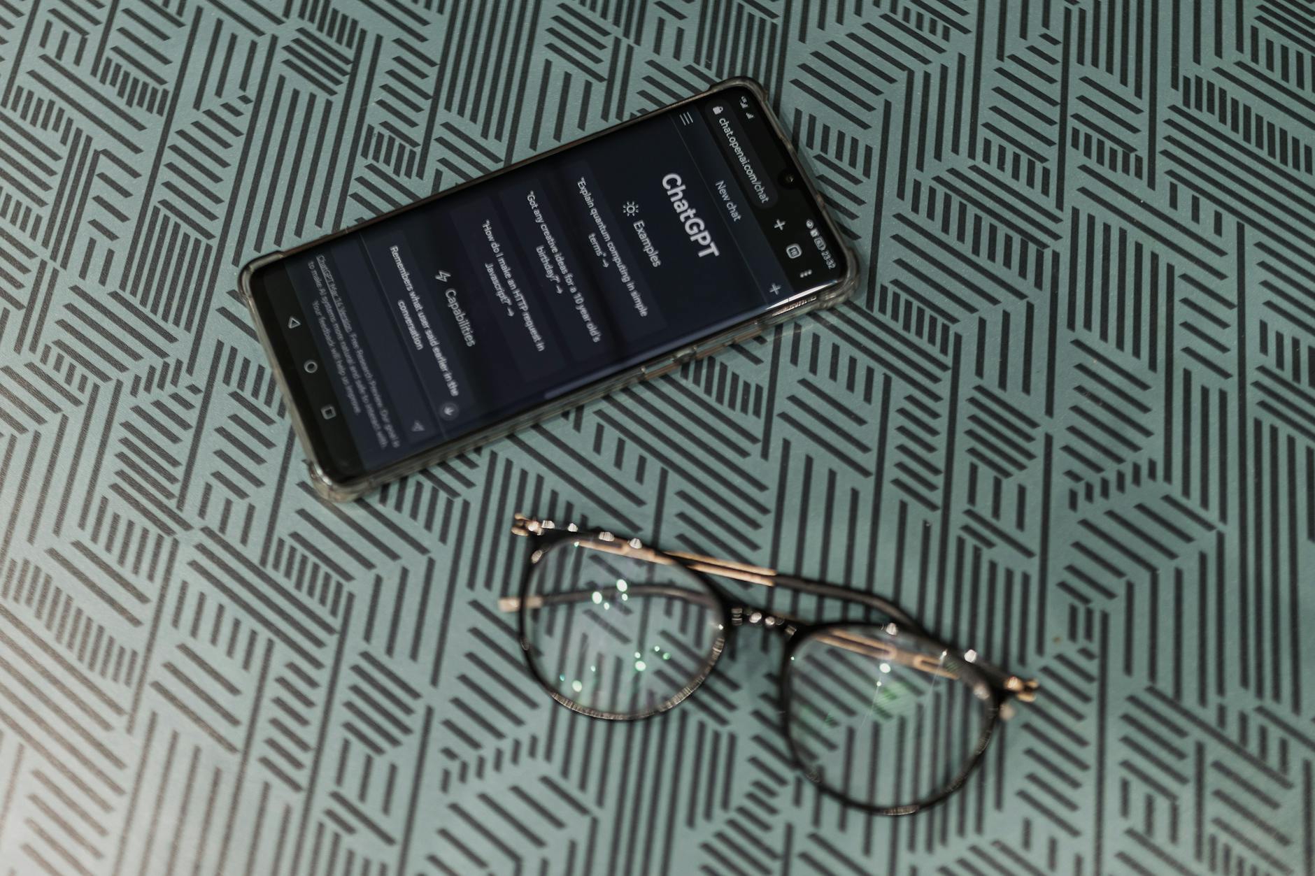 Close-up of smartphone displaying ChatGPT alongside eyeglasses on a patterned surface highlighting technology and style.