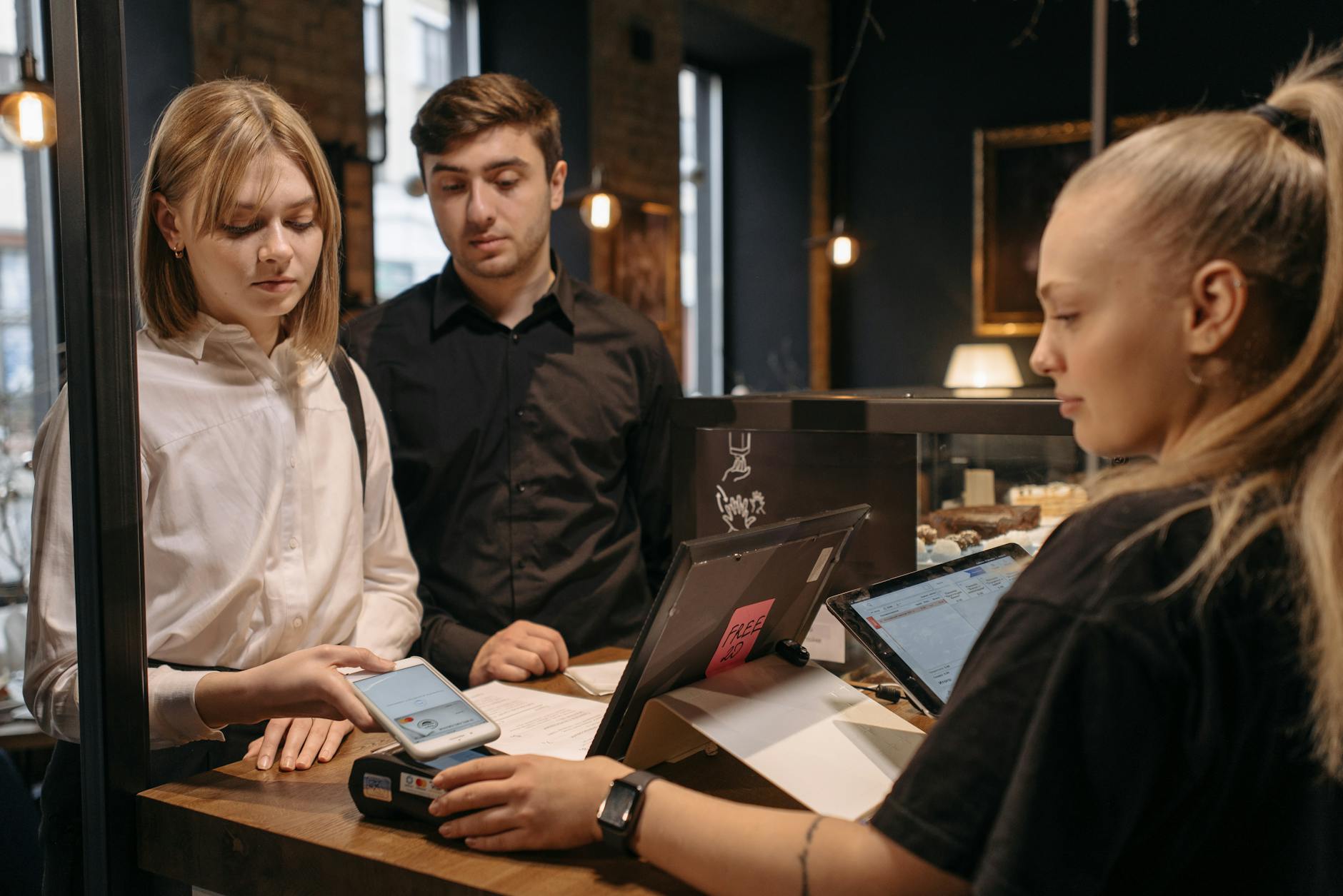 Two customers making a contactless payment at a café counter using a mobile phone.
