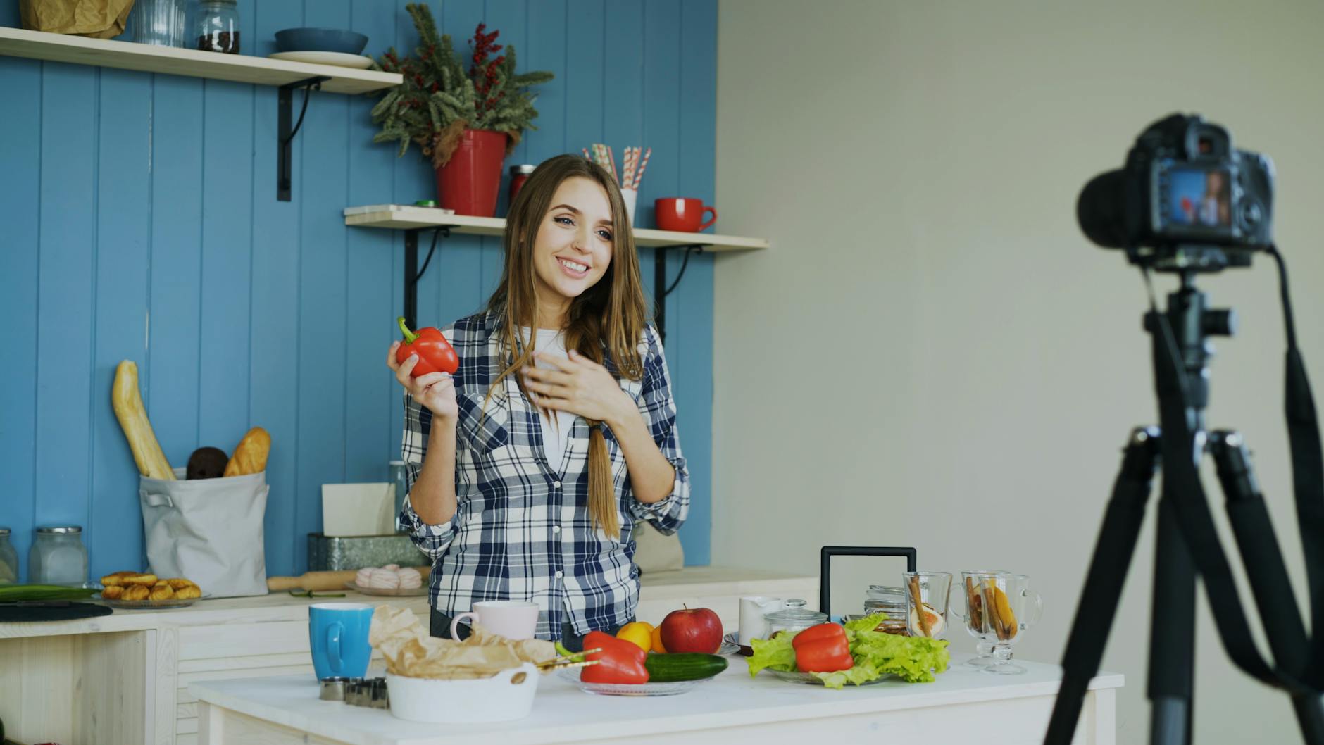 Young woman in a kitchen holding a bell pepper, preparing food, and recording a video.