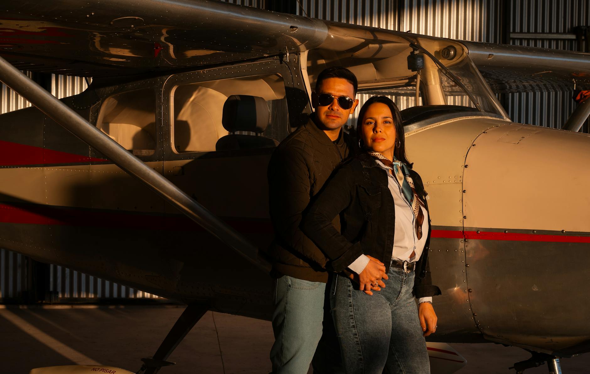 Stylish couple posing by an aircraft inside a hangar during daytime.