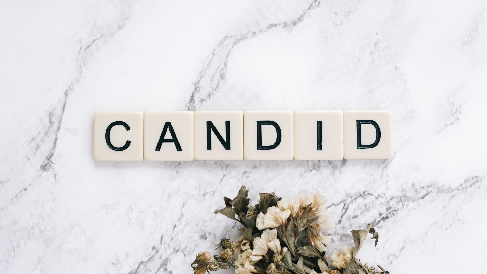 Artistic arrangement of 'CANDID' text tiles on marble surface with dried flowers.