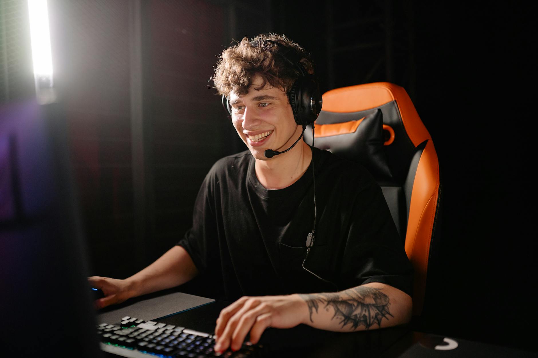 A smiling young man enjoys PC gaming on a computer with headset in an indoor dark setting.