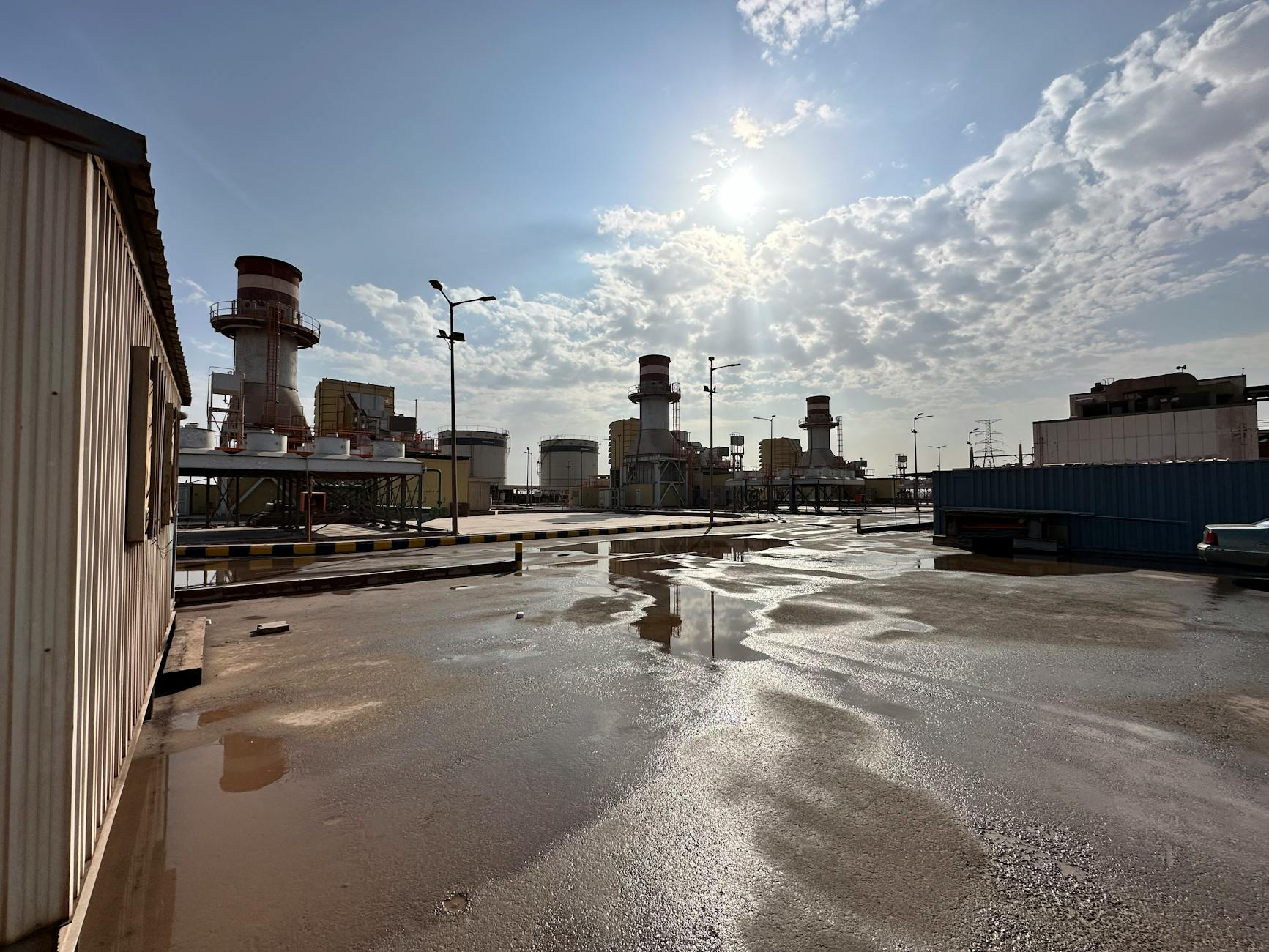 View of an industrial power plant under a cloudy sky with sunlight reflections on wet ground.