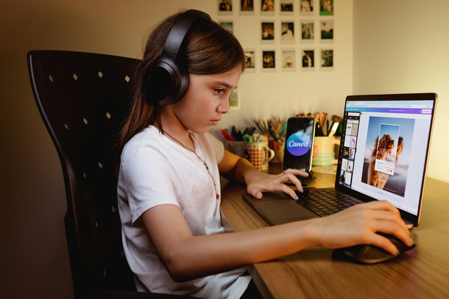 A focused young girl wearing headphones works on a creative project using a laptop and design software.