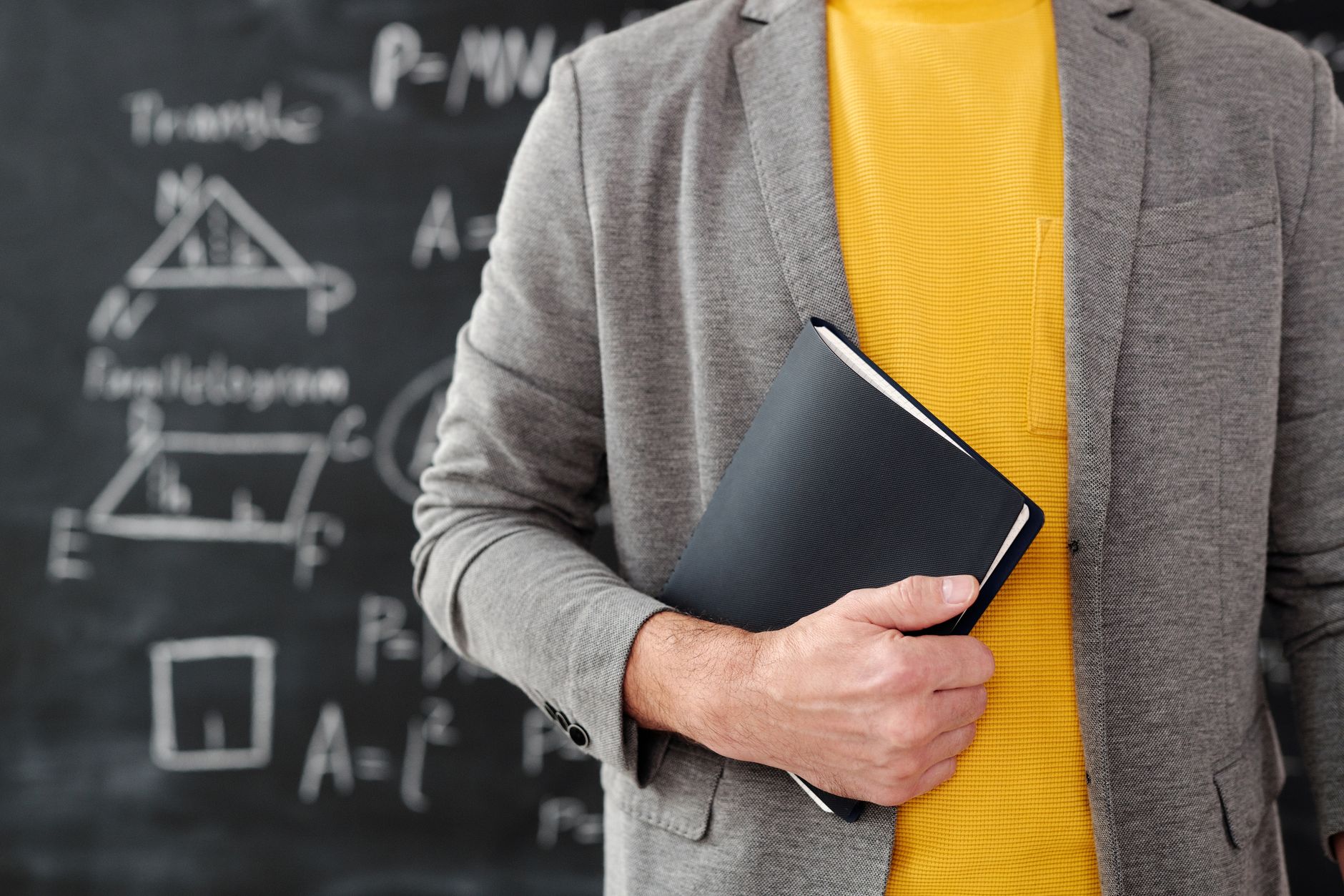 Close-up of a teacher with a notebook standing in front of a blackboard with equations.