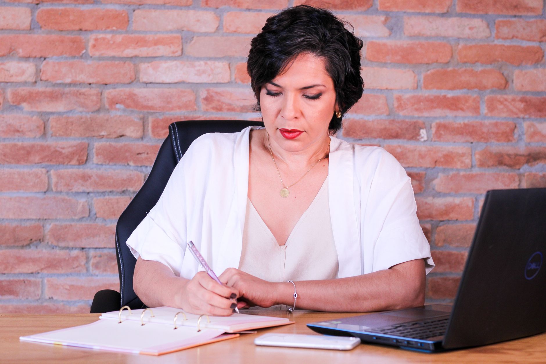 Elegant businesswoman writing in notebook at modern office desk with laptop.